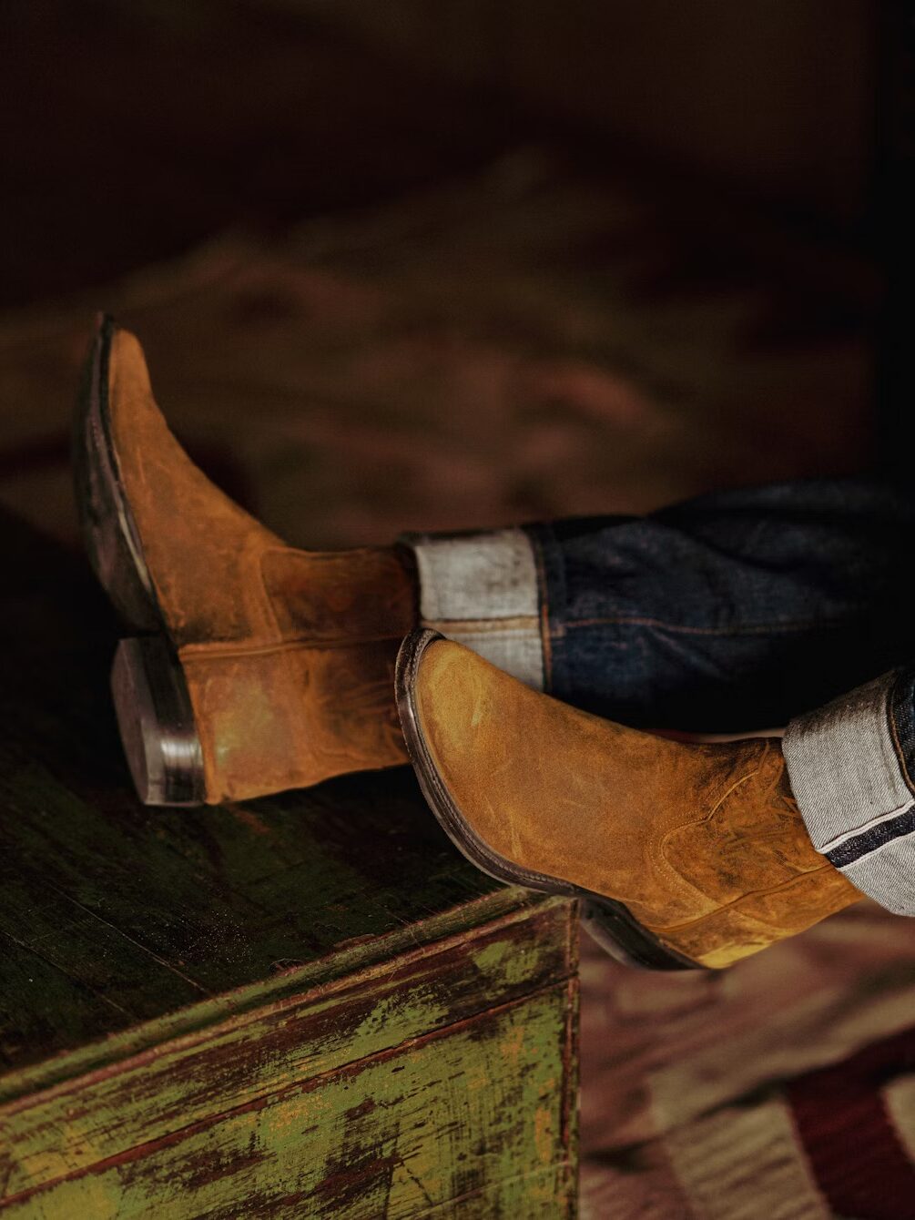 A person wearing rolled-up jeans and brown suede cowboy boots rests their feet on a weathered green wooden surface.