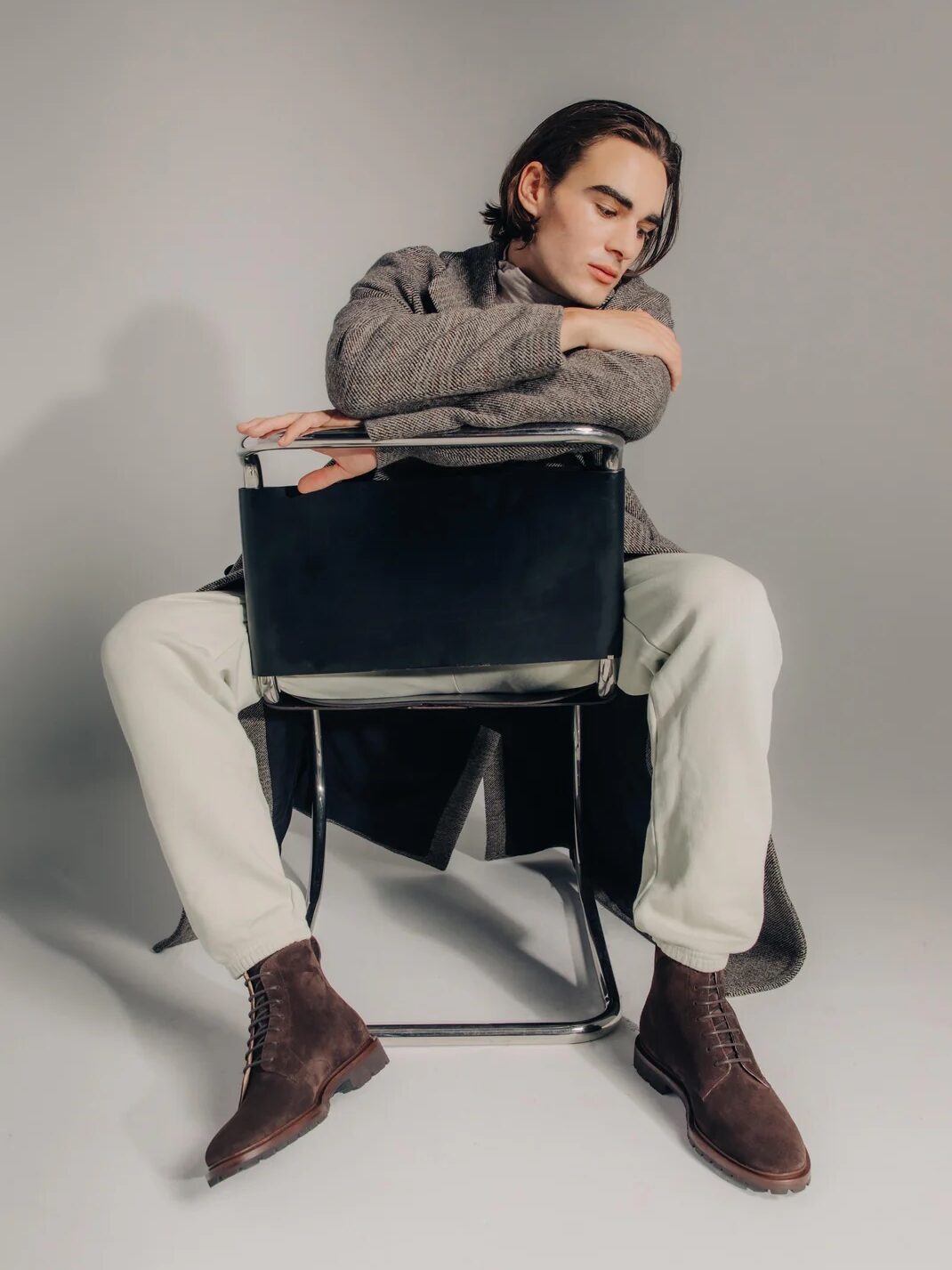A person sits backward on a chair, wearing a gray coat, white pants, and brown boots, with arms resting on the chair back against a plain background.