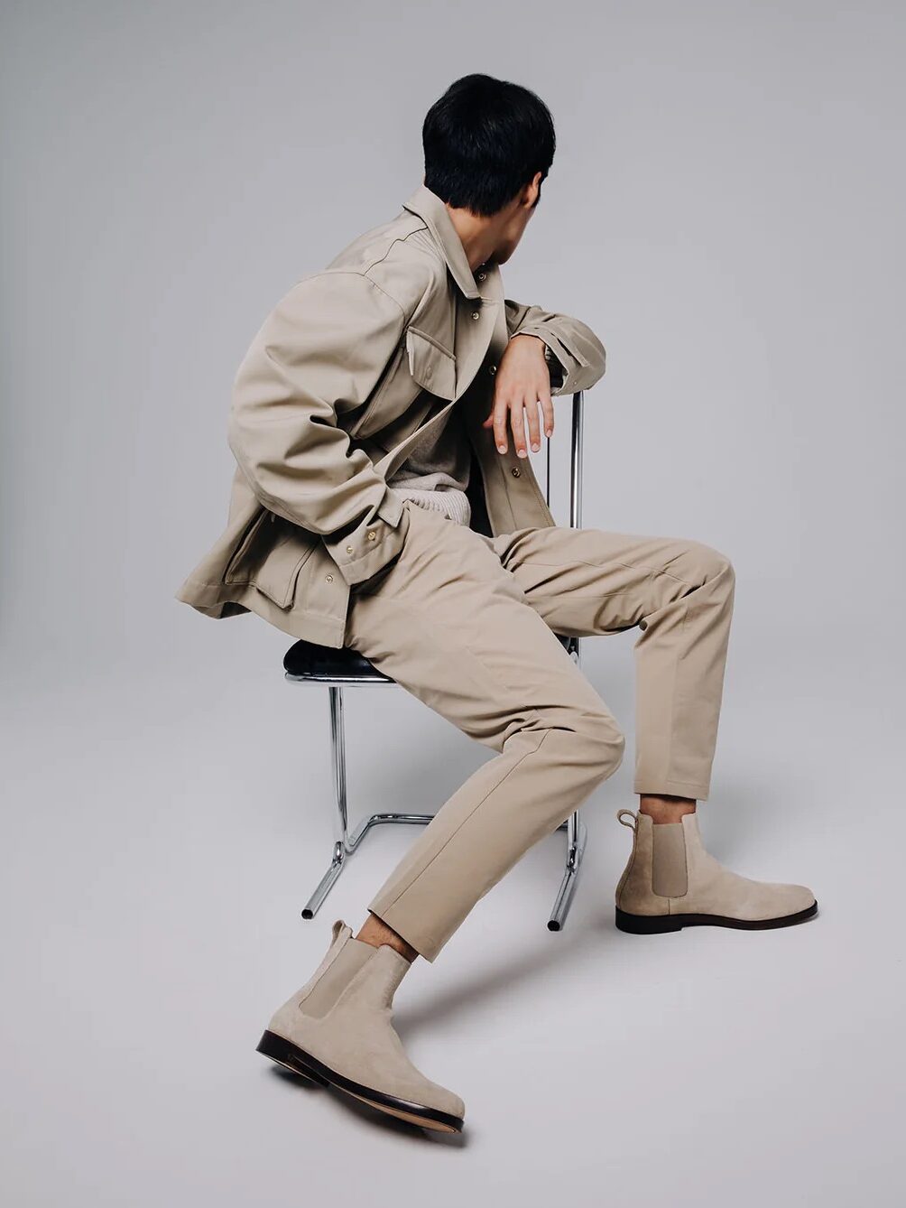 A person in beige clothing and suede Chelsea boots sits on a black chair, facing away in a minimalist studio setting.