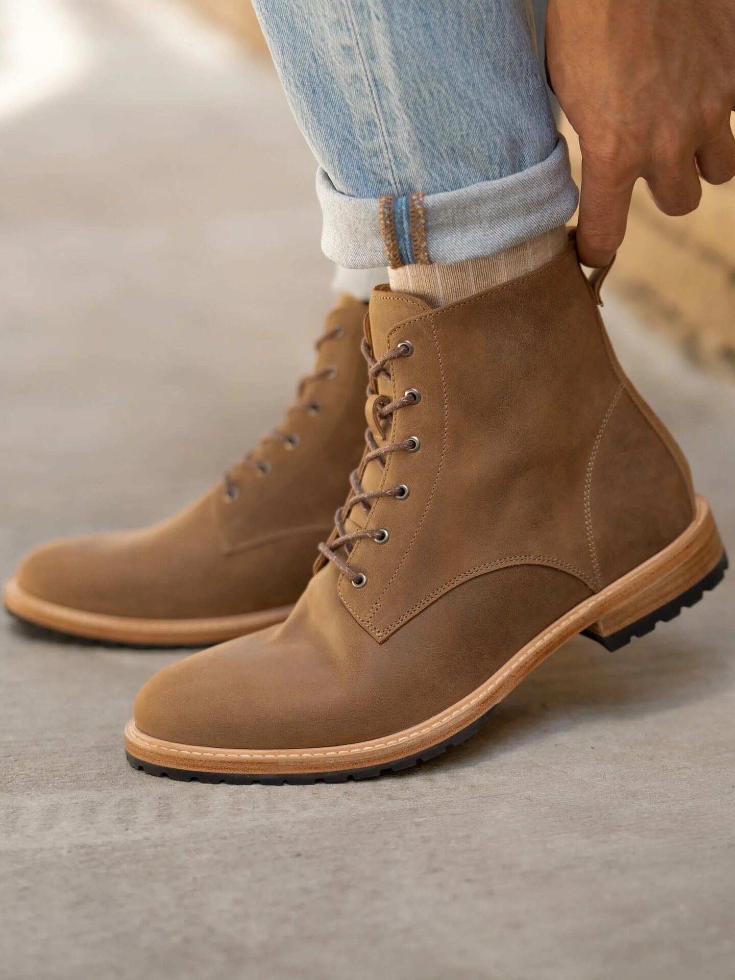 A person wearing light brown leather lace-up boots and light blue cuffed jeans stands on a concrete surface next to a brick wall.