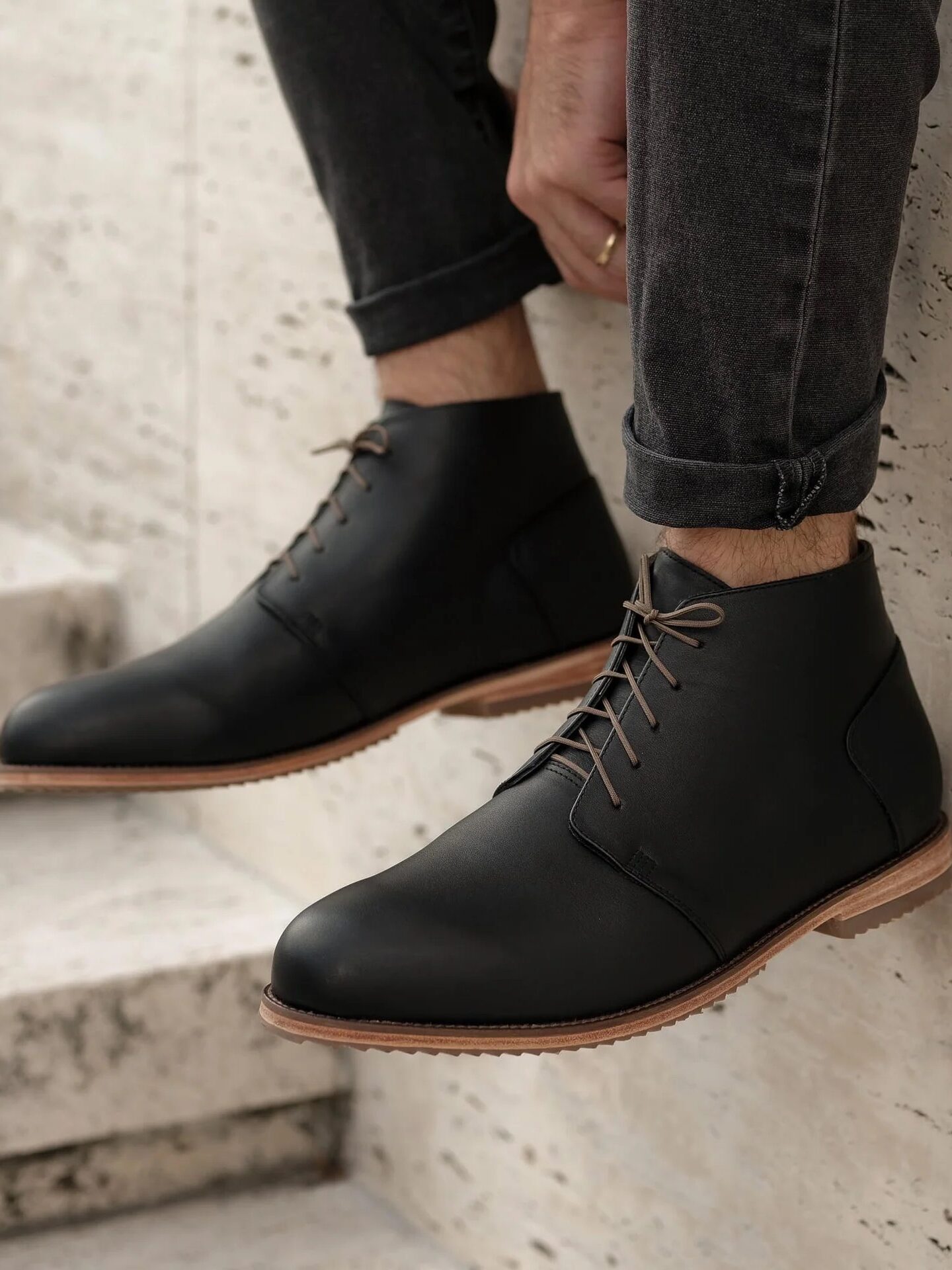 A person wearing black leather lace-up boots and rolled-up dark jeans sits on stone steps next to a textured wall.