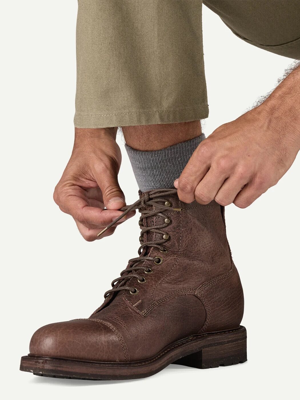 A person tying the laces of a brown leather boot while wearing light brown pants and gray socks.