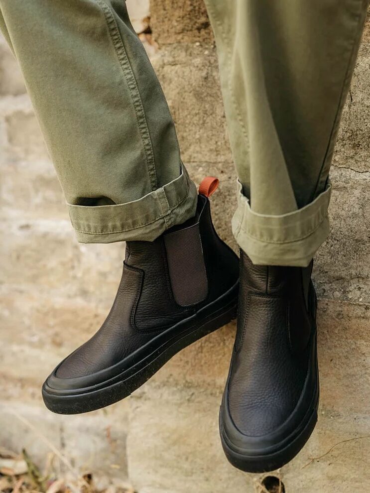 Person wearing olive green pants and black leather Chelsea boots with thick soles, sitting on a stone ledge outdoors.