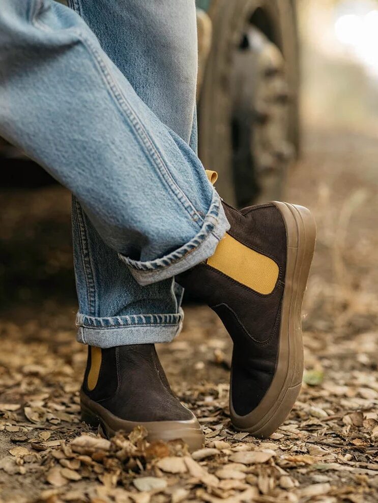 Person wearing cuffed blue jeans and dark brown slip-on boots with yellow elastic sides standing on a leaf-covered dirt path outdoors.