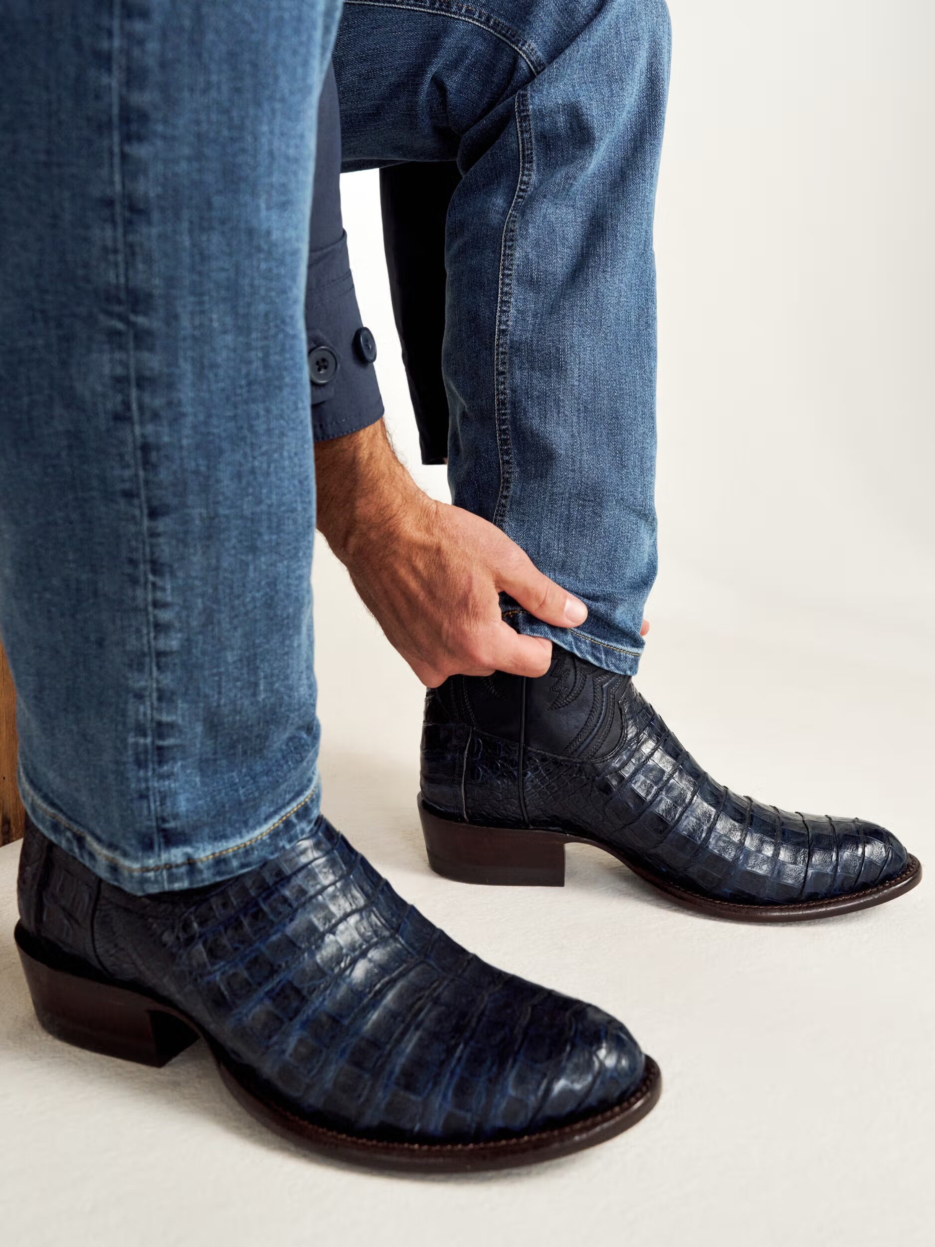A person wearing blue jeans adjusts the cuff of their pant leg over a pair of dark blue, crocodile-patterned leather boots.