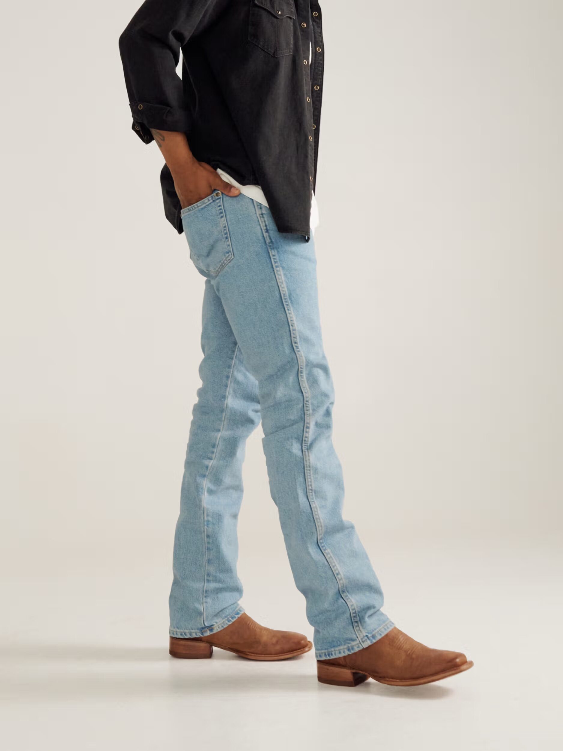 Person wearing a black button-up shirt, light blue jeans, and brown cowboy boots stands with hands in pockets against a plain background.