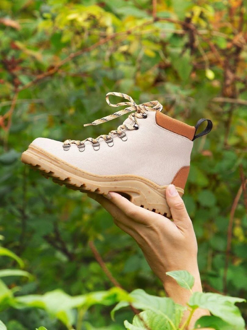 A hand holds up a tan hiking boot with brown accents and patterned laces against a green, leafy outdoor background.