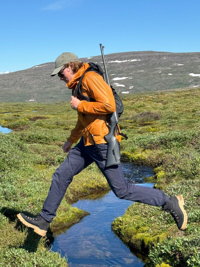 Person wearing outdoor gear and a backpack jumps over a narrow stream in a grassy, mountainous landscape under a clear blue sky.