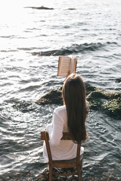 A person with long hair sits on a wooden chair at the edge of the water, reading a book with sunlight reflecting off the sea.