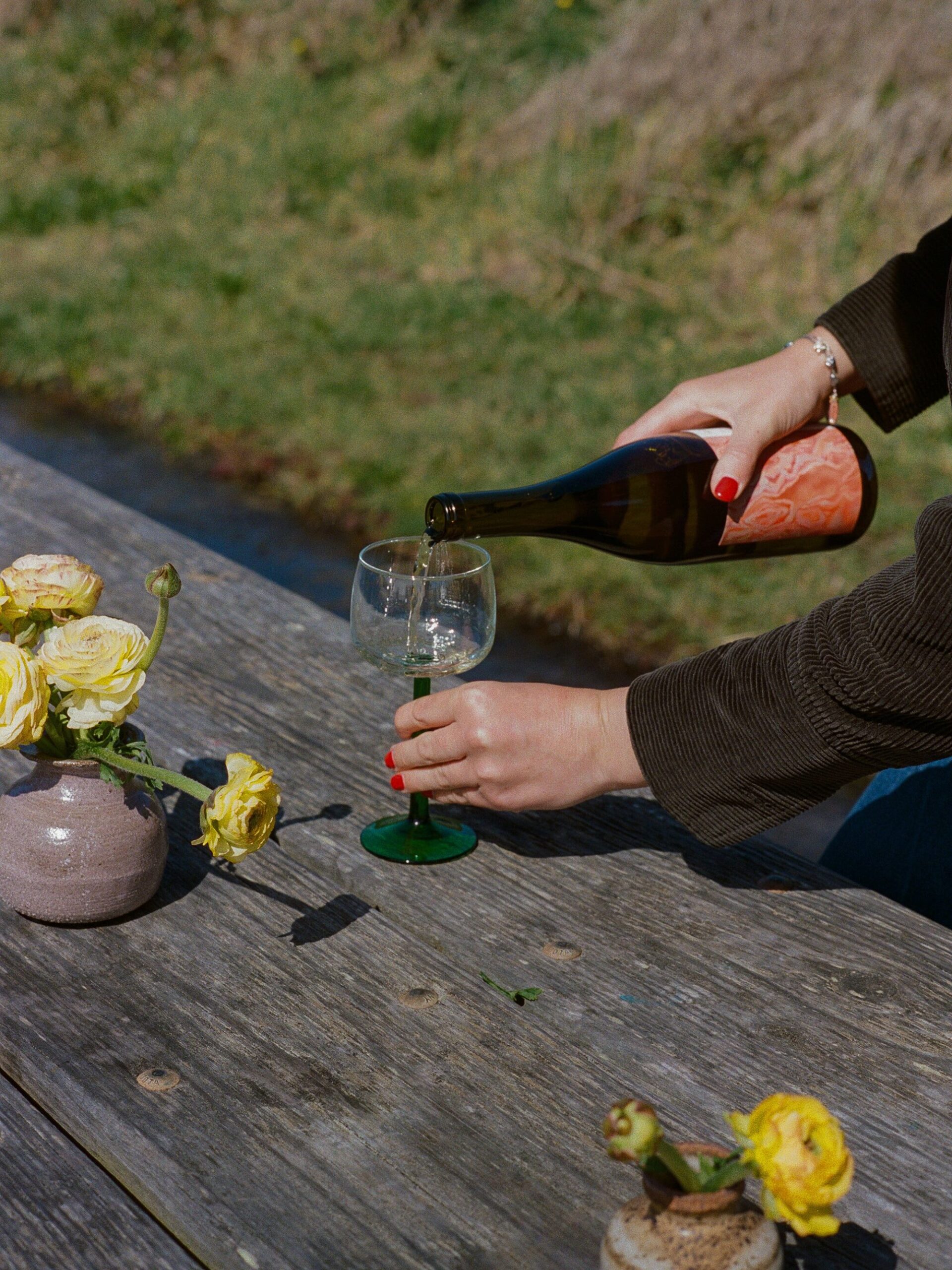 A person pours white wine into a glass on a wooden outdoor table, with yellow flowers in small vases nearby.