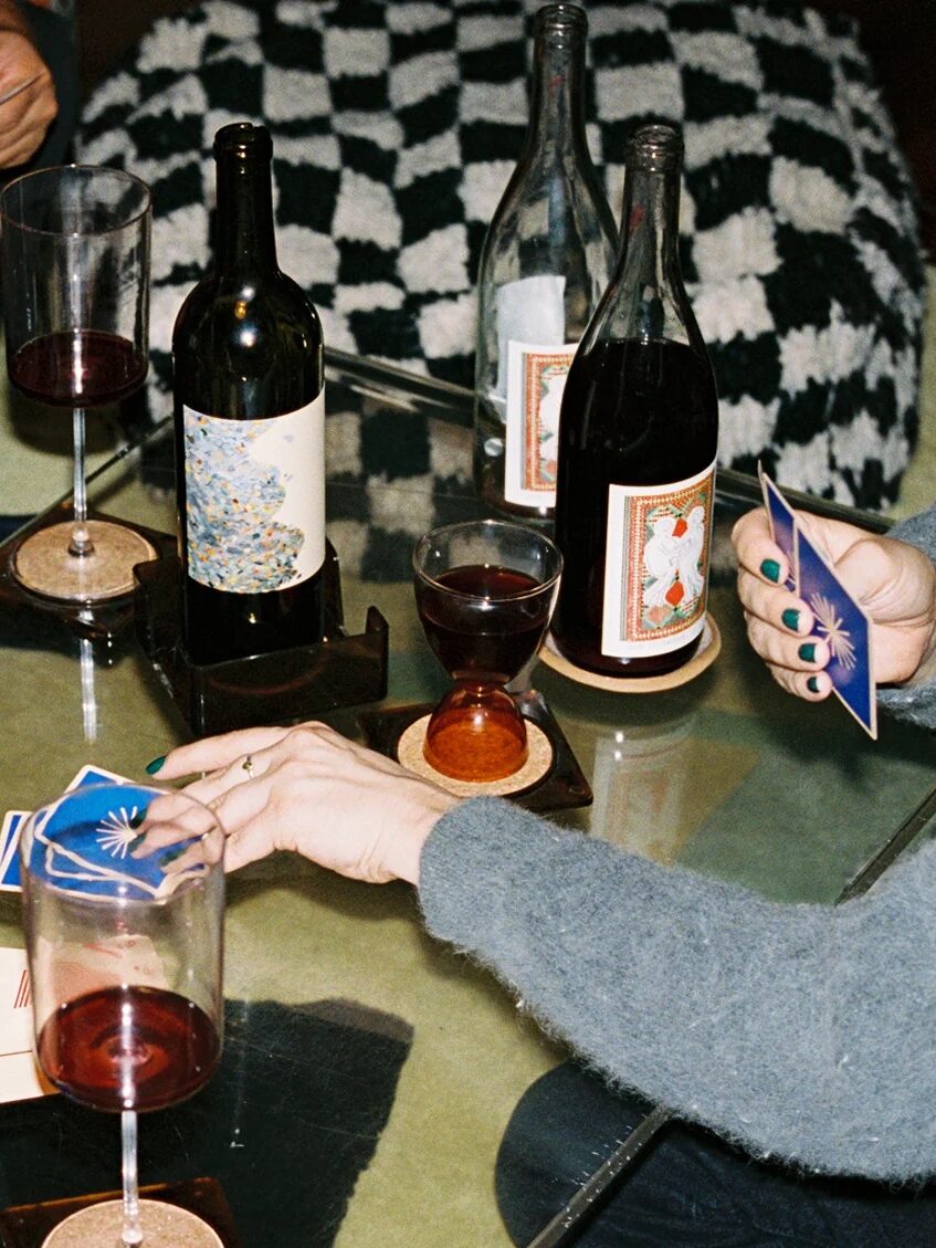 Four people sit around a glass table playing a card game, with several glasses of red wine and three wine bottles visible on the table.