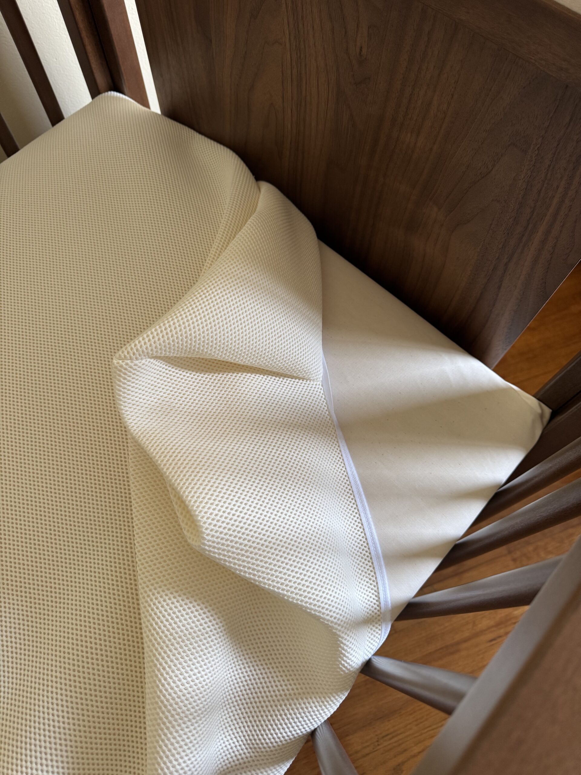 A close-up of a crib mattress with a textured fitted sheet partially pulled back, revealing a smooth, white mattress cover underneath.