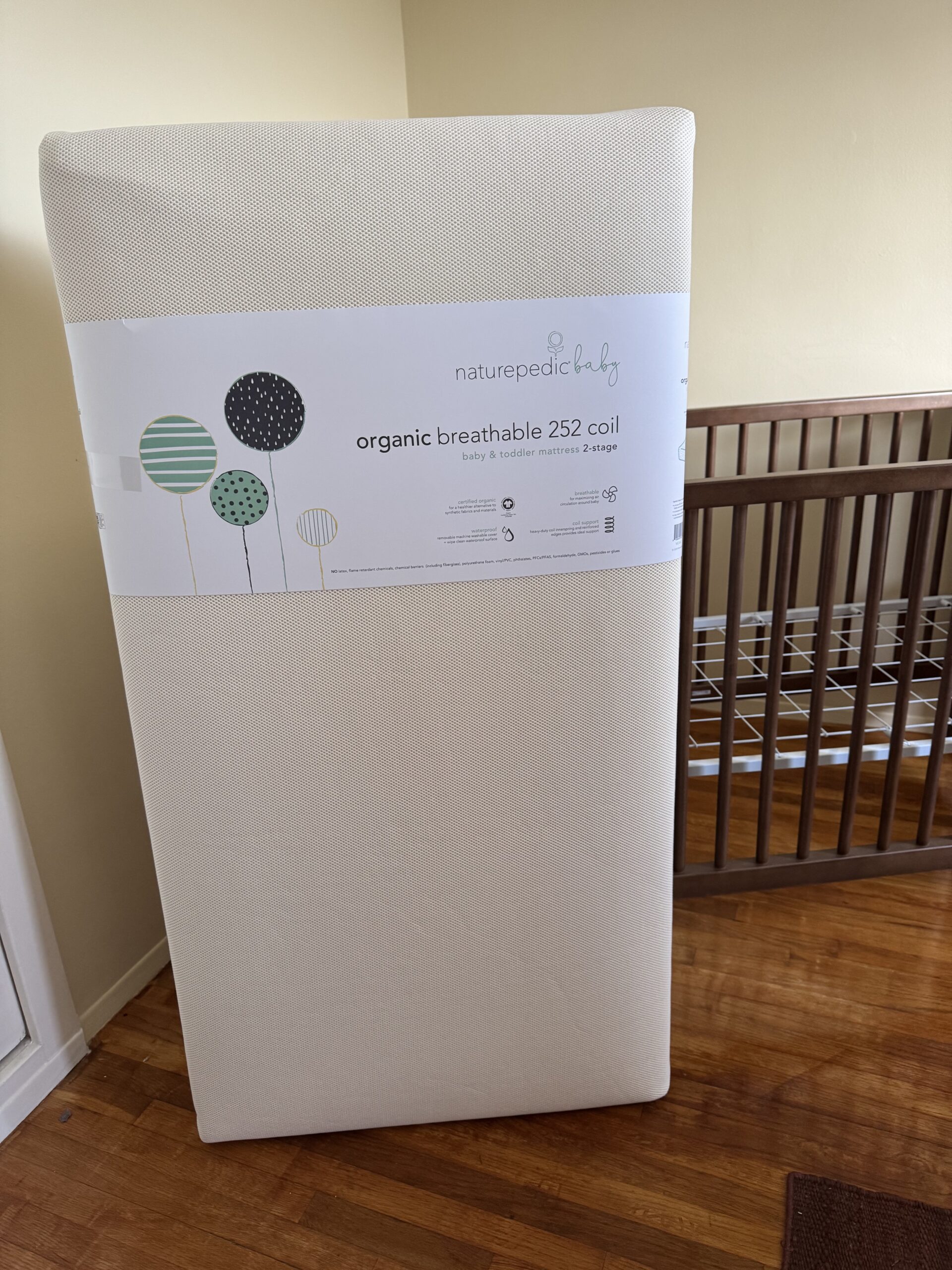 A Naturepedic baby crib mattress labeled "organic breathable 252 coil" stands upright next to a wooden crib on a hardwood floor.