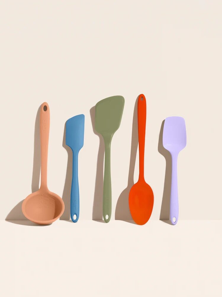 Five silicone kitchen utensils in pastel and bold colors, including a ladle, two spatulas, a spoon, and a scraper, are arranged upright against a neutral background.