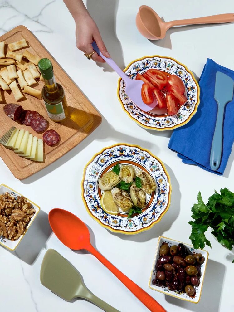 A hand serves sliced tomatoes from a decorative plate. Nearby are a cutting board with cheese, meat, and oil, bowls of walnuts and olives, spatulas, a ladle, herbs, and a folded napkin.