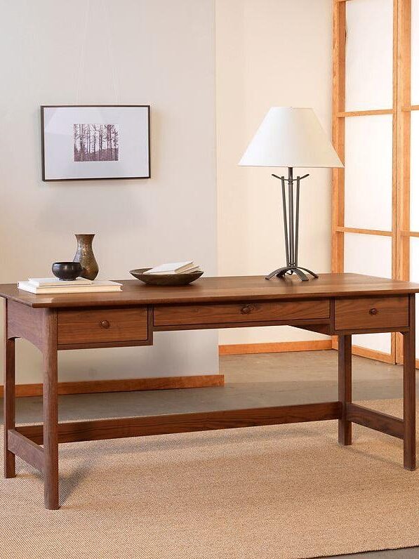 Wooden desk with a small drawer, accompanied by a lamp, bowls, and a vase. A framed picture is on the wall. A potted plant and dresser are in the background.