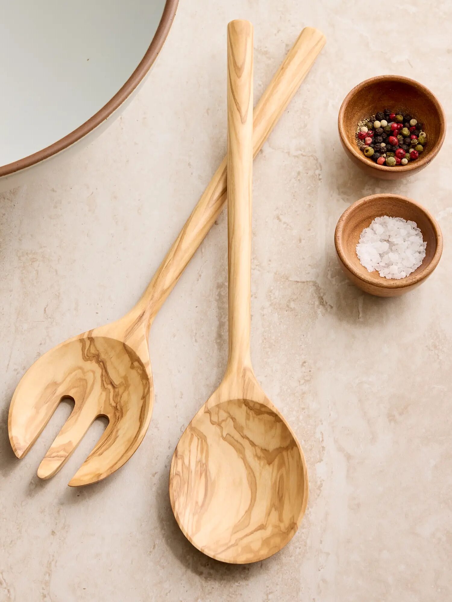 Two wooden salad servers are crossed on a countertop next to small bowls containing peppercorns and coarse salt, with part of a large bowl visible in the corner.