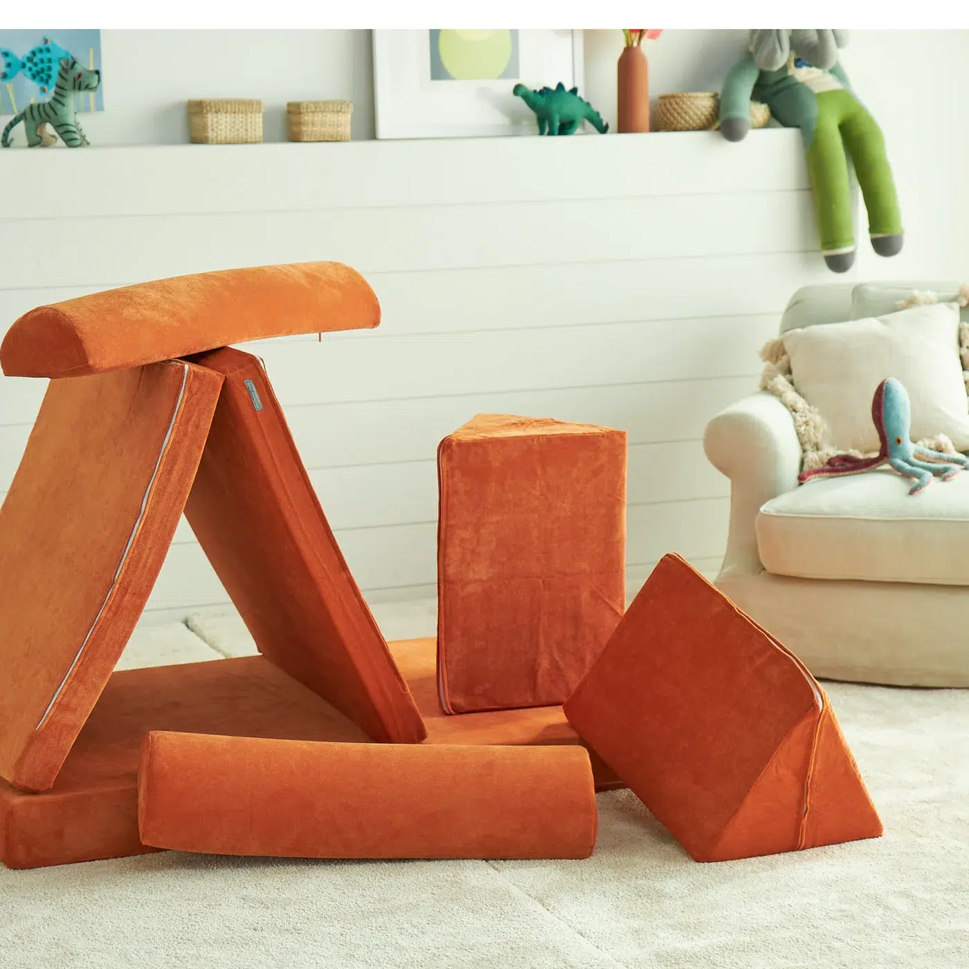 Orange foam play couch pieces are arranged in a fort shape on a carpeted floor, with toys and an armchair in the background.