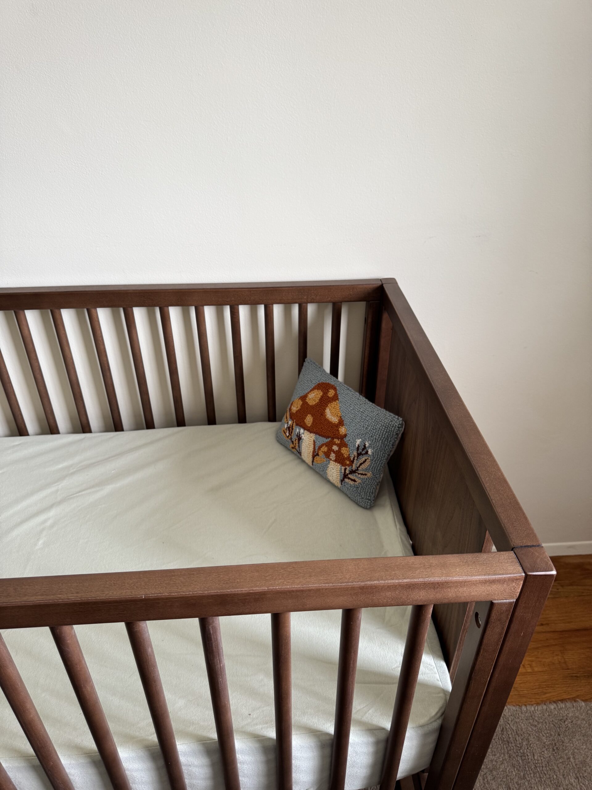 A wooden crib with a white mattress and a pillow featuring an orange deer design, positioned against a plain white wall.