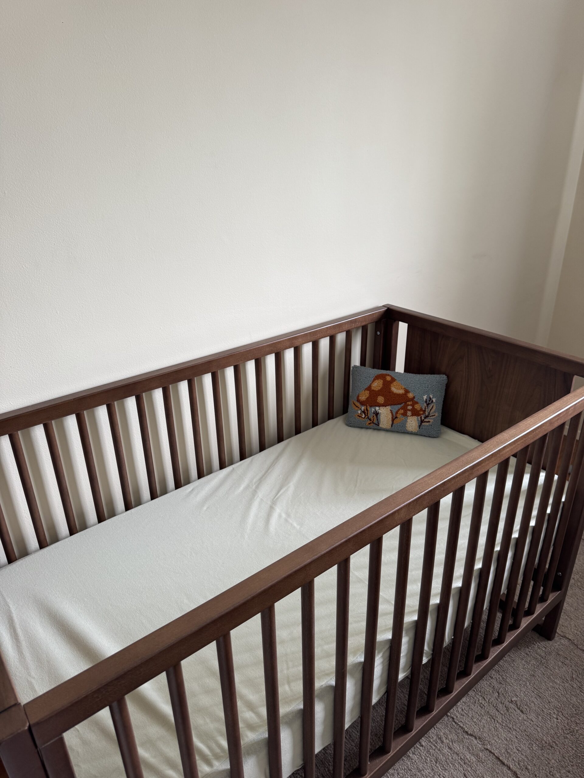 A wooden baby crib with a white mattress and a decorative pillow featuring a mushroom and deer, placed against a plain light-colored wall.