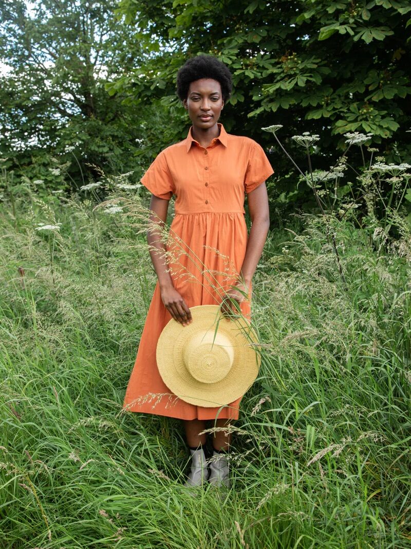 Person in an orange dress stands in tall grass, holding a yellow hat, with green foliage in the background.