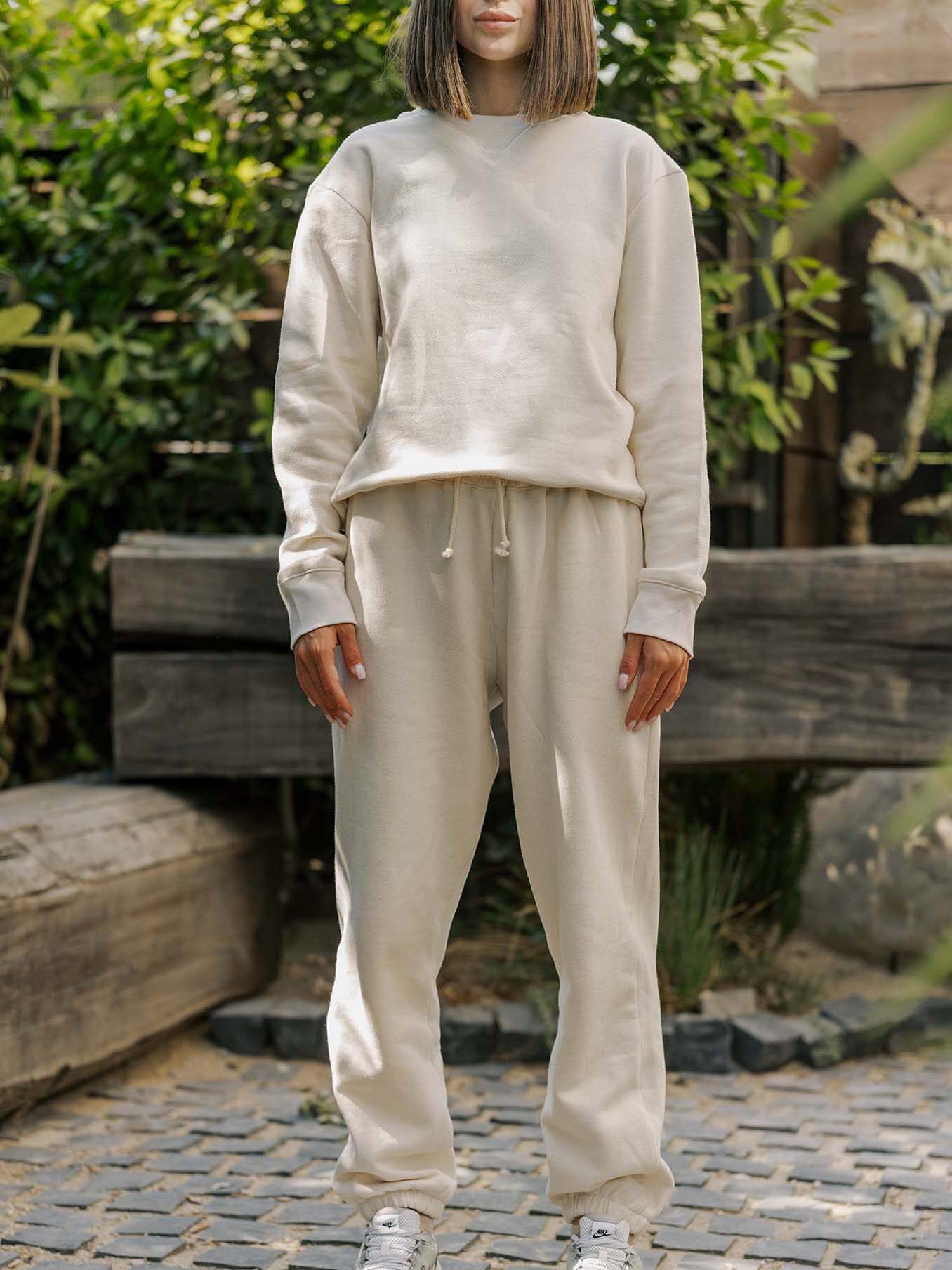Person standing outdoors wearing a beige sweatshirt and sweatpants with white sneakers, set against a background of greenery and cobblestone pavement.