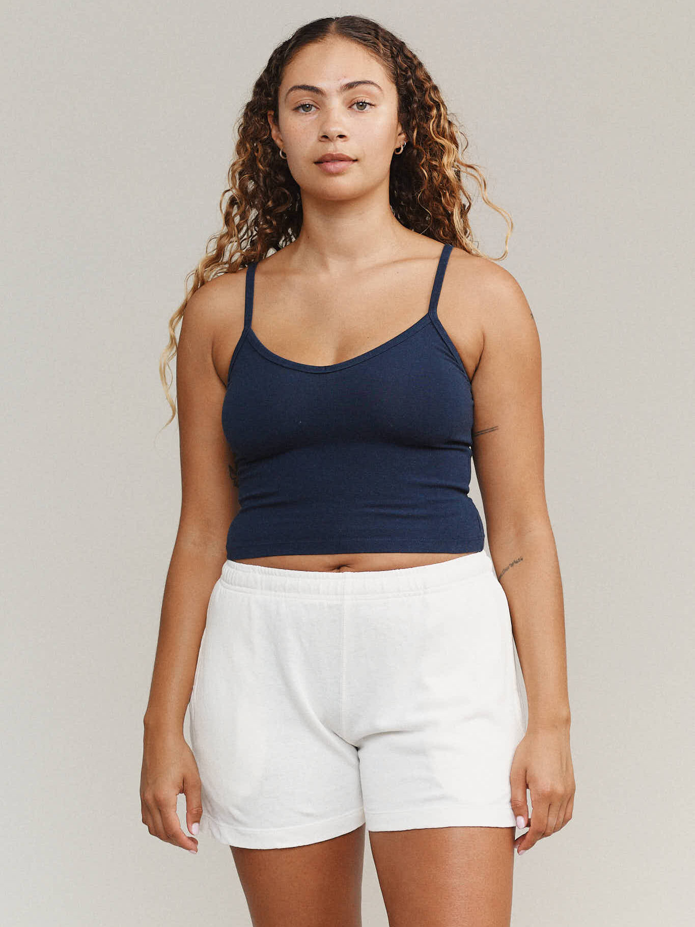 A person with long curly hair wearing a navy tank top and white shorts stands against a plain background.