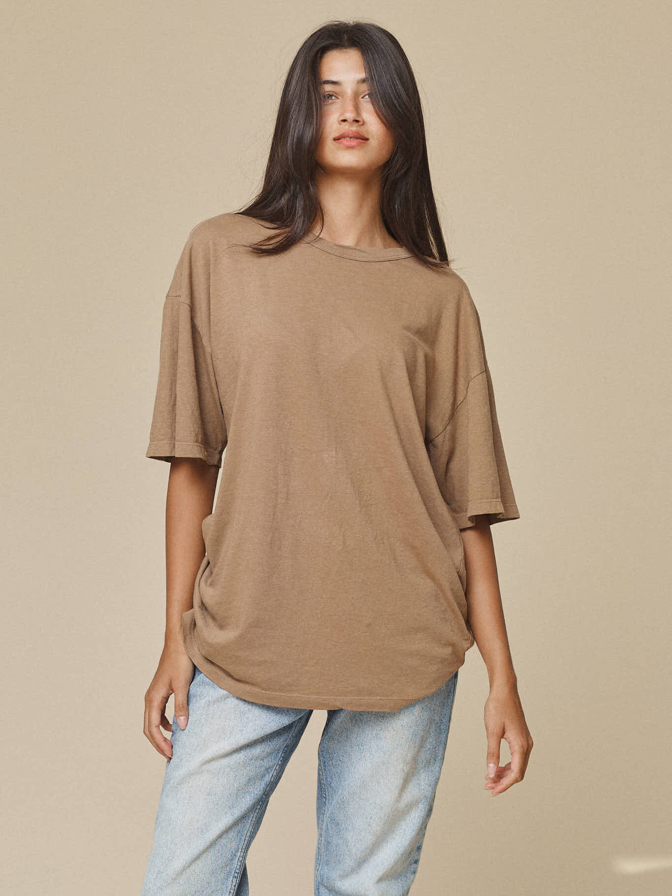 Person wearing a loose-fitting brown t-shirt and light blue jeans stands against a plain beige background.