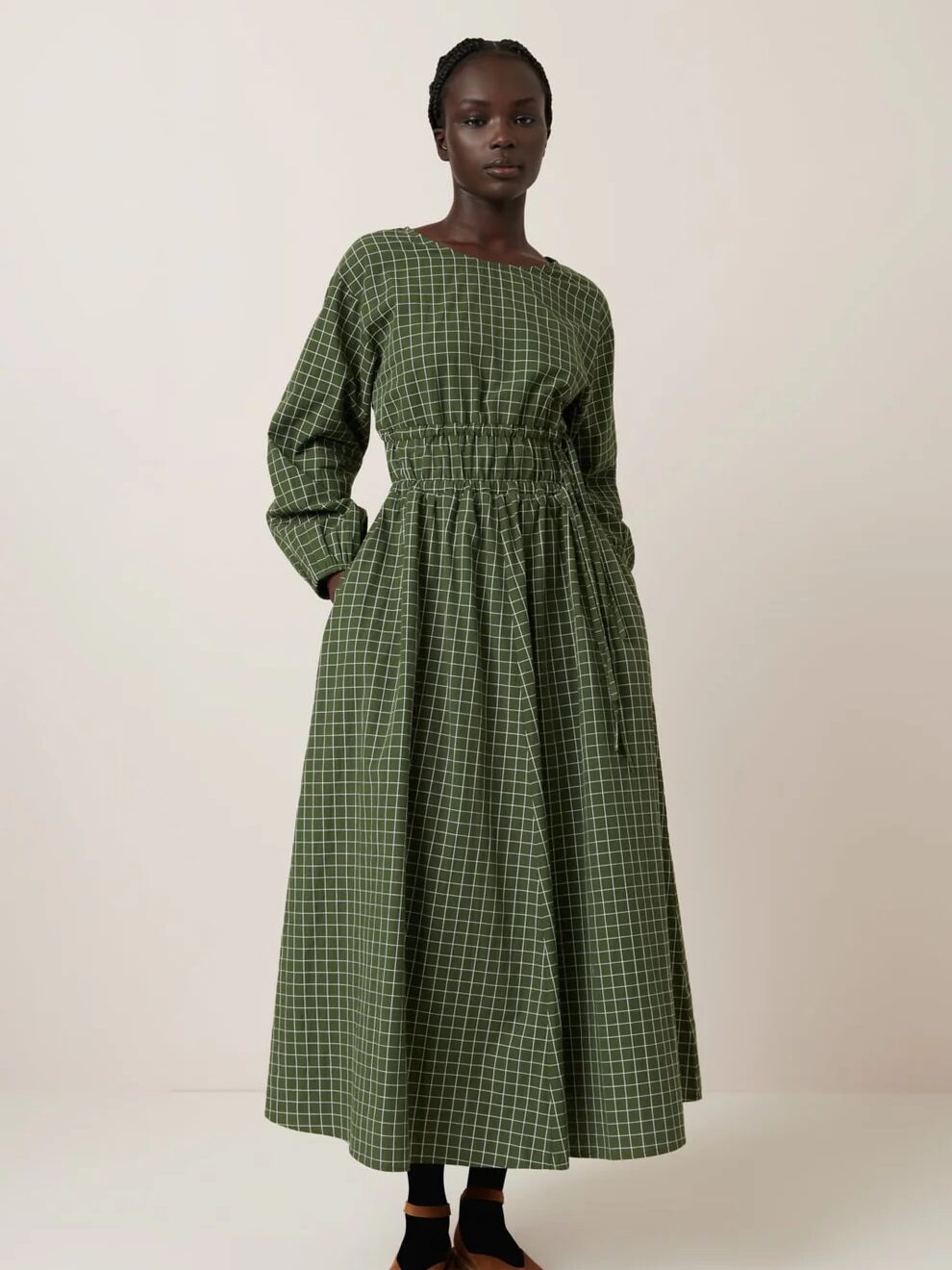 Person wearing a long-sleeved, green checkered dress with a cinched waist, black socks, and brown shoes, standing against a plain background.