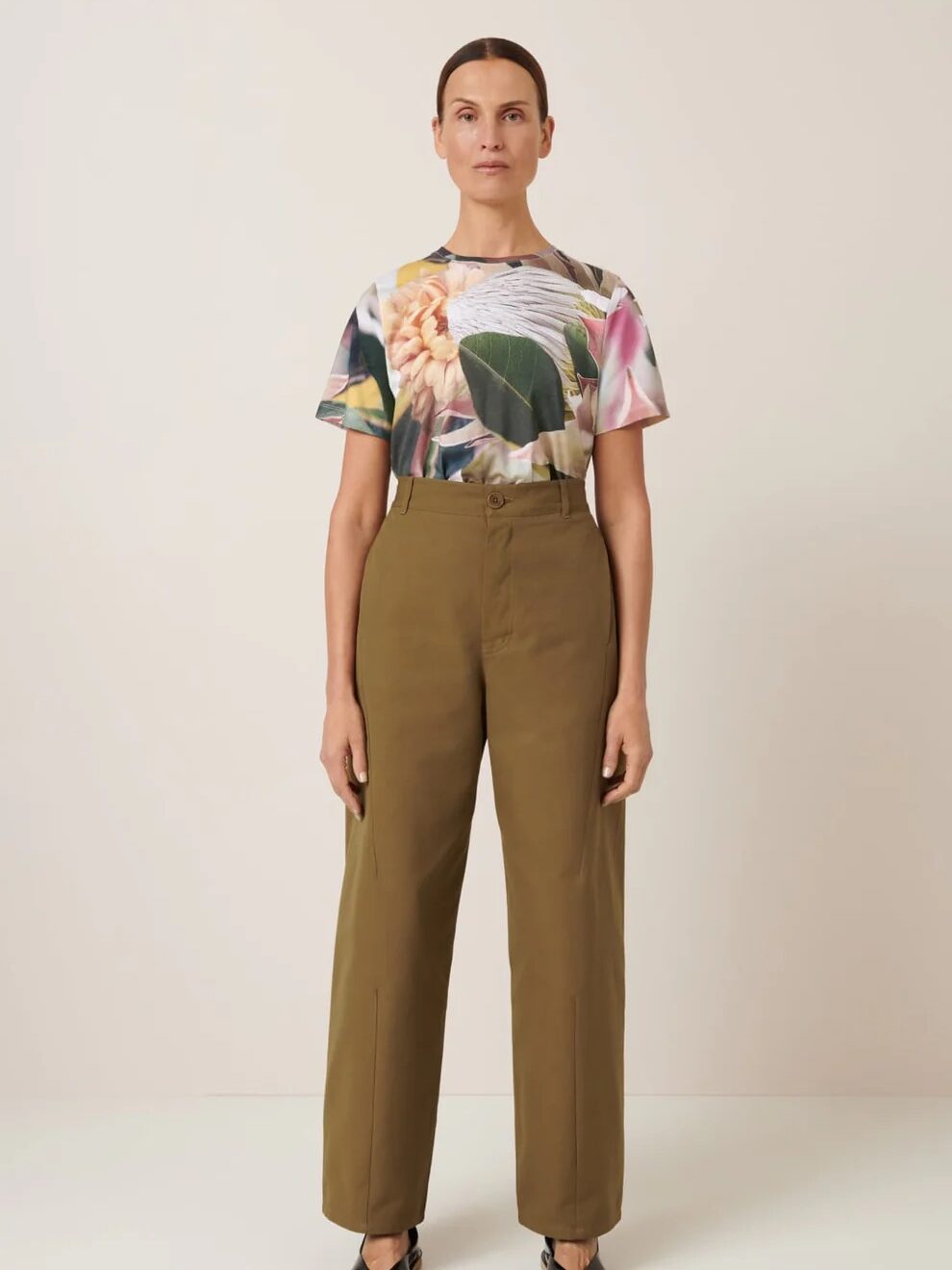 Person standing against a neutral background wearing a floral print shirt and high-waisted, olive green trousers, with black open-toe shoes.