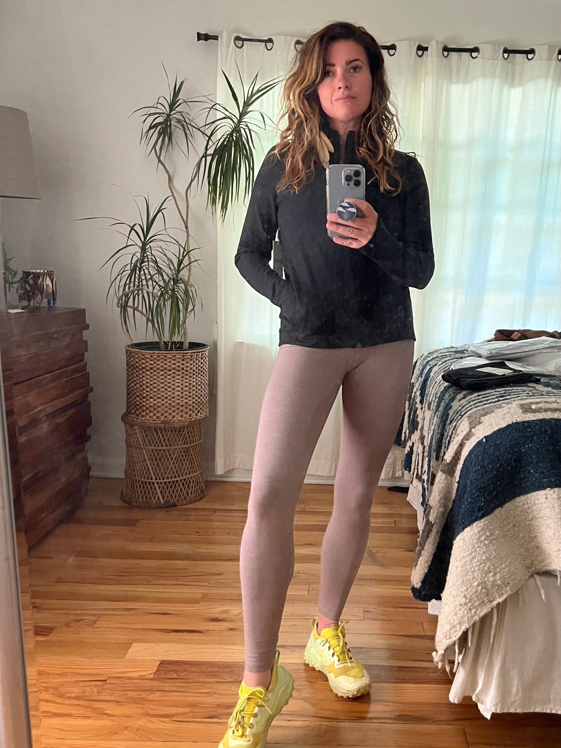 A person in a black long-sleeve shirt, beige leggings, and yellow sneakers takes a mirror selfie in a room with plants, a bed, and a lamp.