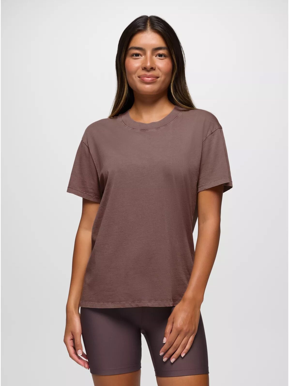 A person wearing a brown t-shirt and shorts stands against a neutral background, smiling with hands at their sides.