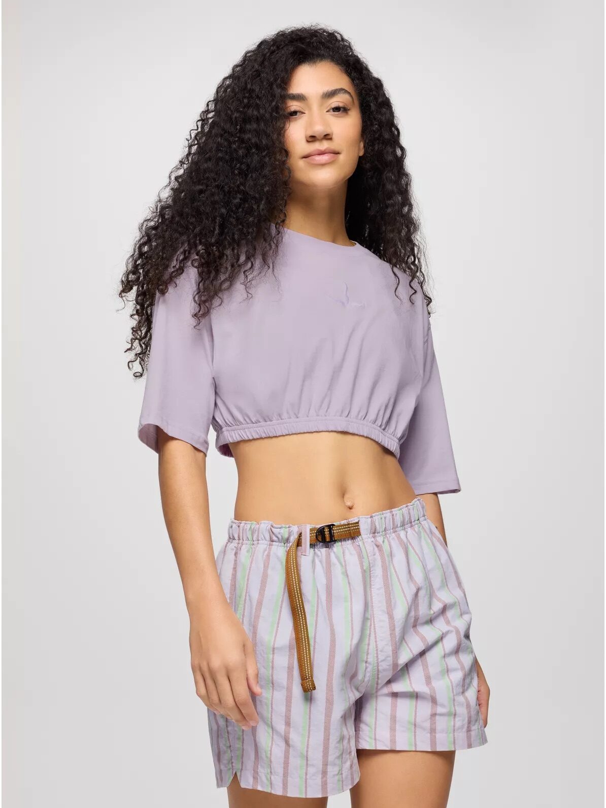 A person with long curly hair wearing a light purple crop top and striped shorts, standing against a plain background.
