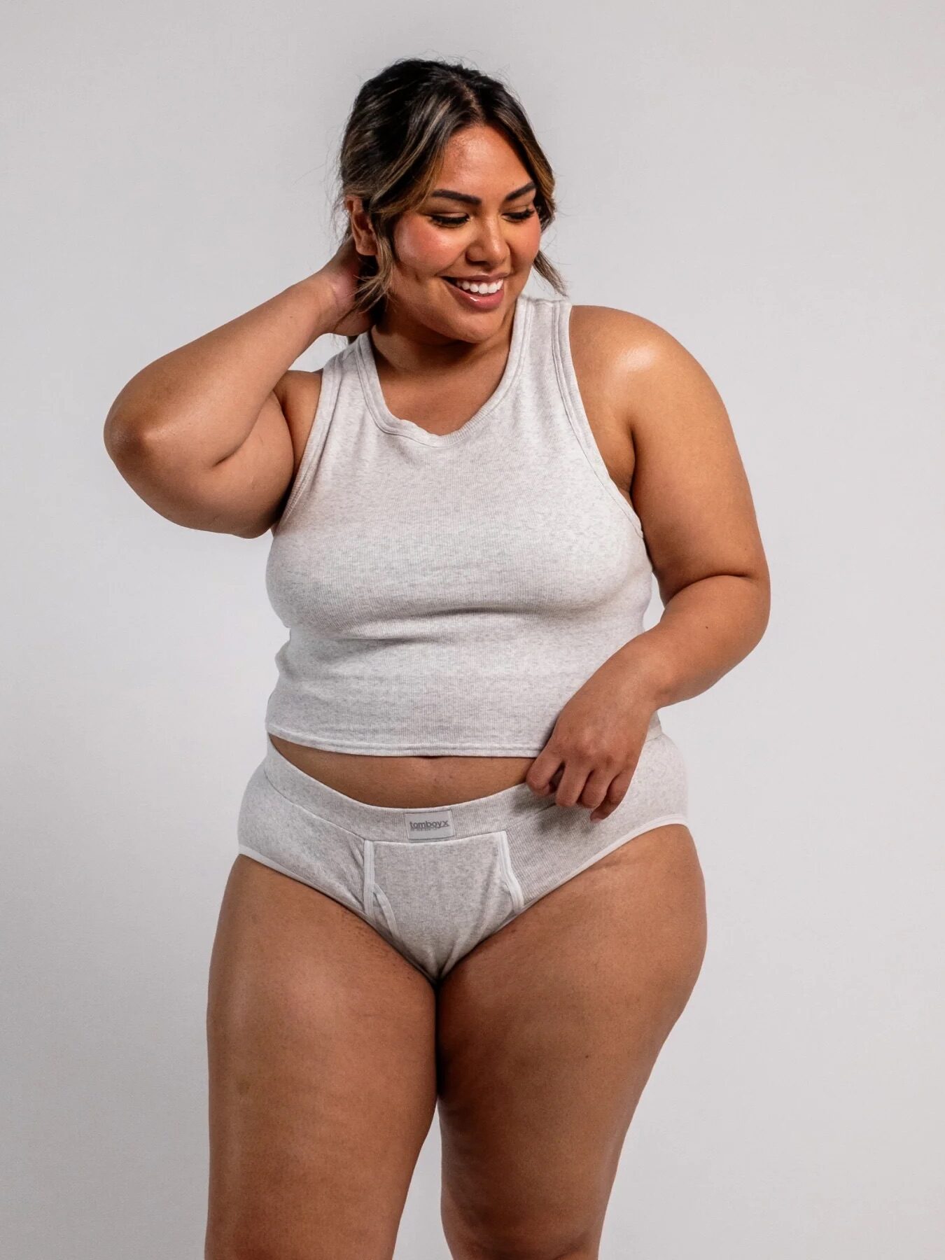 Woman in a gray tank top and underwear stands smiling, with one hand on her neck against a plain background.