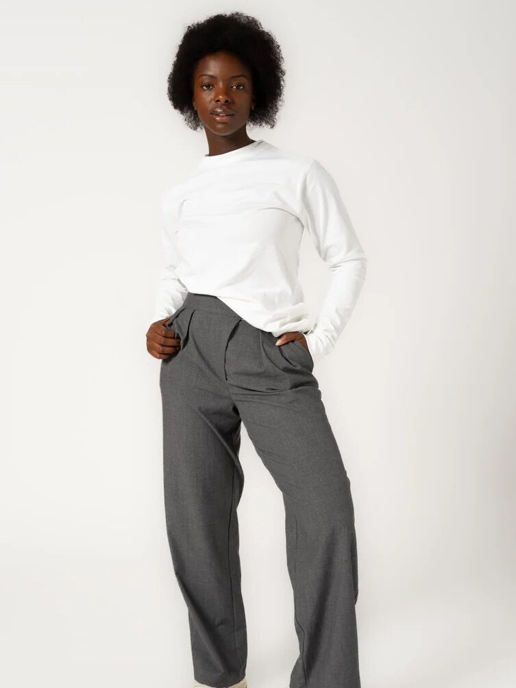 Person wearing a white long-sleeve shirt, gray pants, and beige shoes stands against a plain white background.