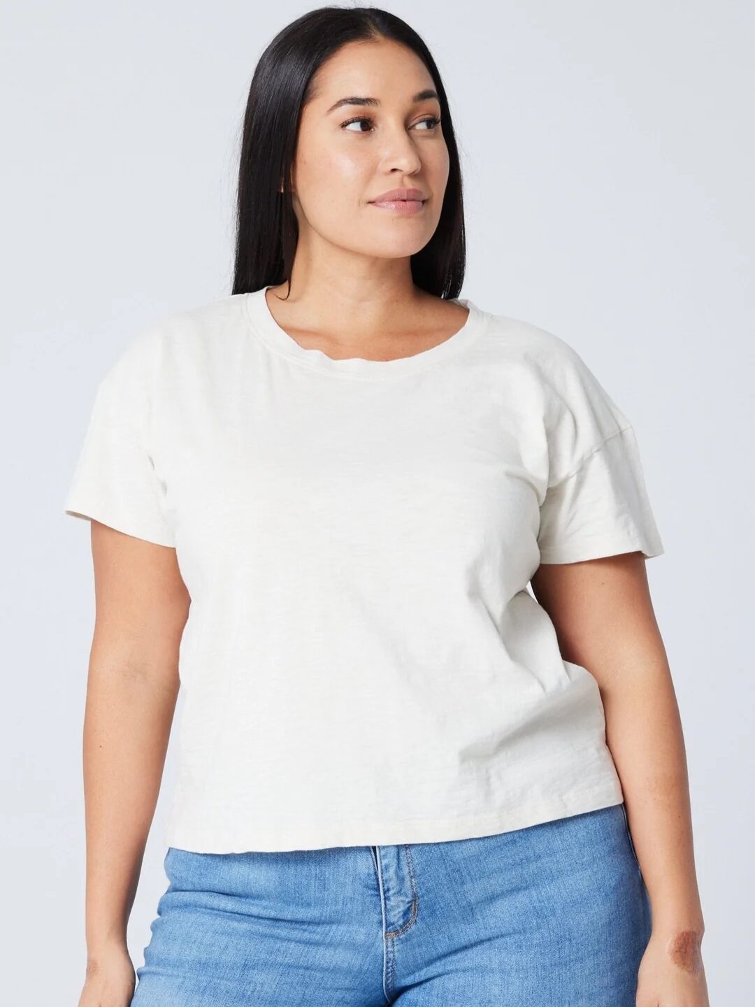 A woman with long dark hair wears a plain white short-sleeve t-shirt and blue jeans, standing against a light background.