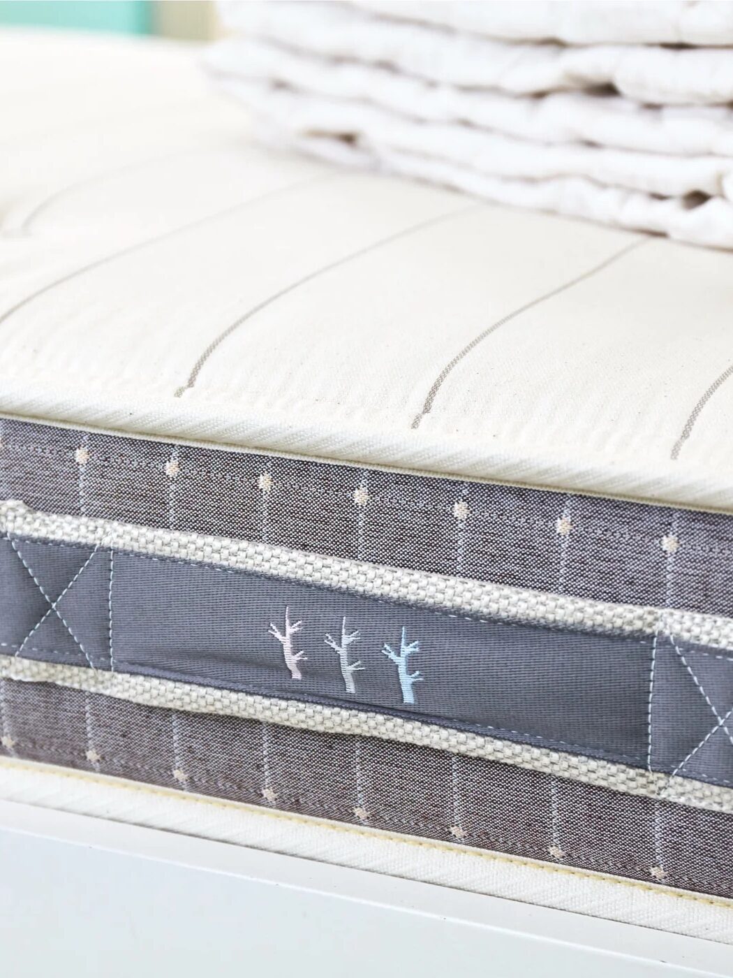 Close-up of a mattress with a striped pattern and a handle on the side, featuring embroidered minimalist tree designs. Folded bedding is stacked on top.