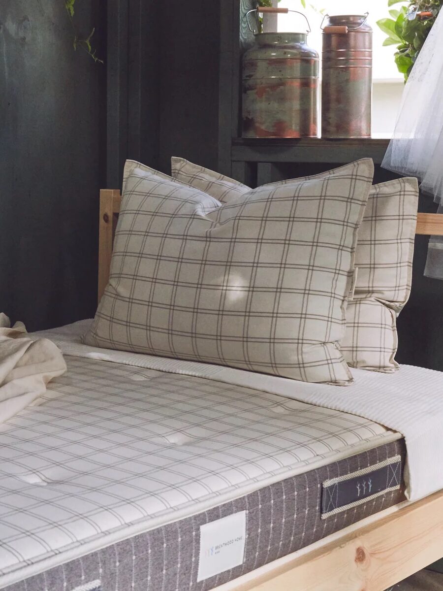 A rustic bed with plaid-patterned bedding, pillows, and a light blanket, situated near a window with green foliage outside. Copper containers are placed on a shelf above the bed.