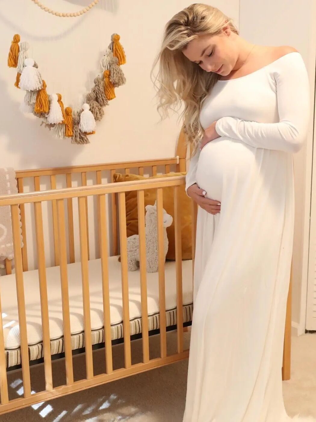 Pregnant woman in a white dress stands by a wooden crib in a nursery, touching her belly. Decorative tassels hang above the crib.