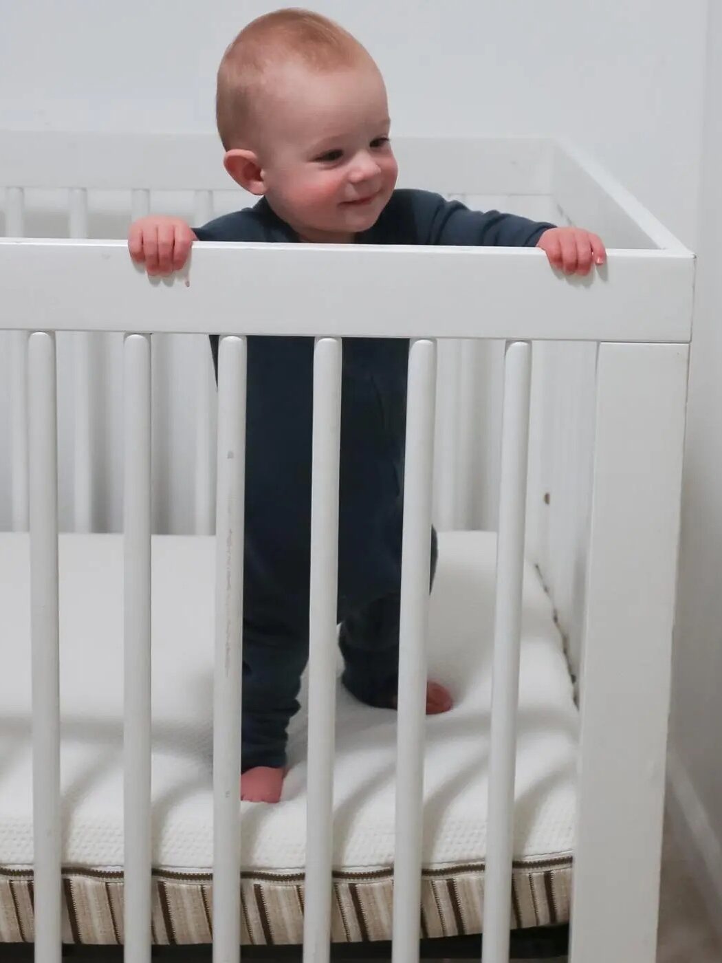 Baby standing and holding the side of a white crib, wearing a dark onesie, smiling.