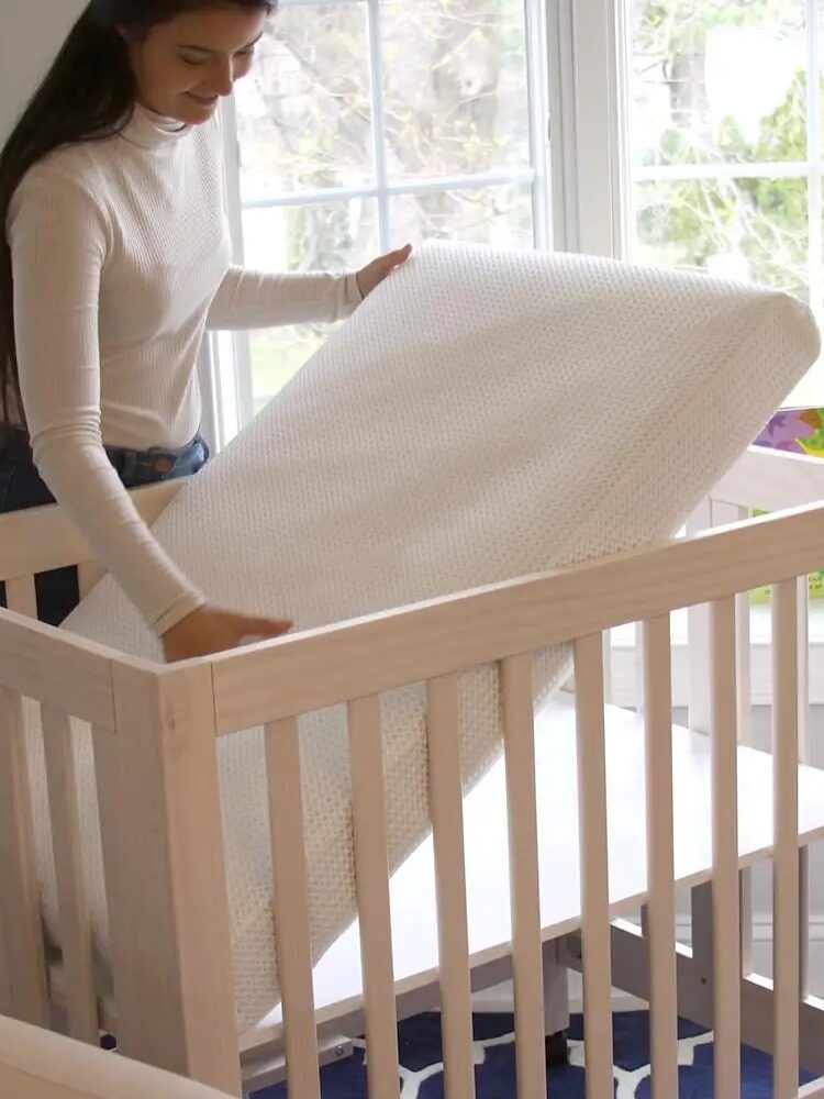 A person places a white mattress into a wooden crib near a window in a bright room.