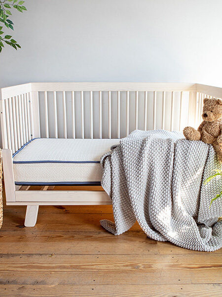A wooden crib with a gray blanket and teddy bear beside a potted fern. A wicker basket with pillows is nearby on a wooden floor with a plant in the corner.