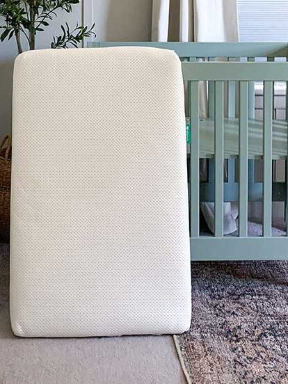 Foam mattress stands upright in a nursery with a green crib, a rug, and a small table with a giraffe toy.