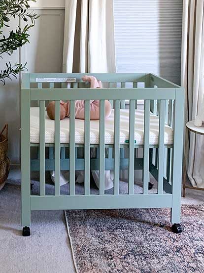 A pastel green crib with a baby inside is in a nursery. The room features a potted plant, a small table with plush toys, and a hanging garment. Light curtains cover the window.