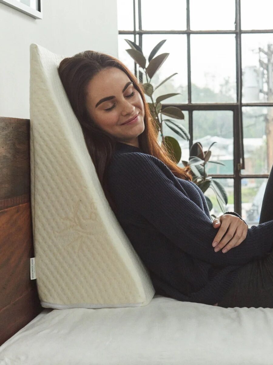 Woman sits on a bed, leaning comfortably against a beige wedge pillow with a textured cover, near a large window with plants in the background.