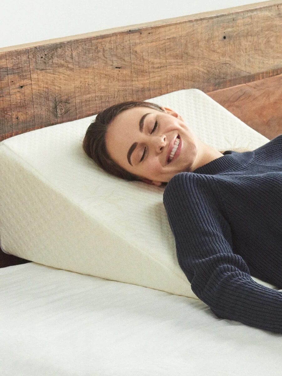 A woman lies on a bed, smiling with her eyes closed, propped up by a large wedge pillow against a wooden headboard.