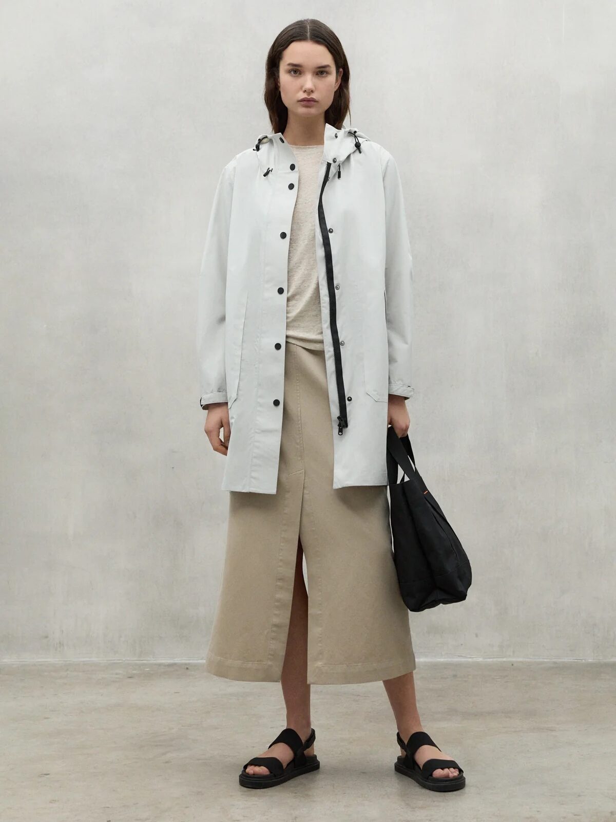 A woman stands indoors wearing a light-colored waterproof raincoat over a beige skirt and top, holding a black bag and wearing black sandals against a plain gray wall.