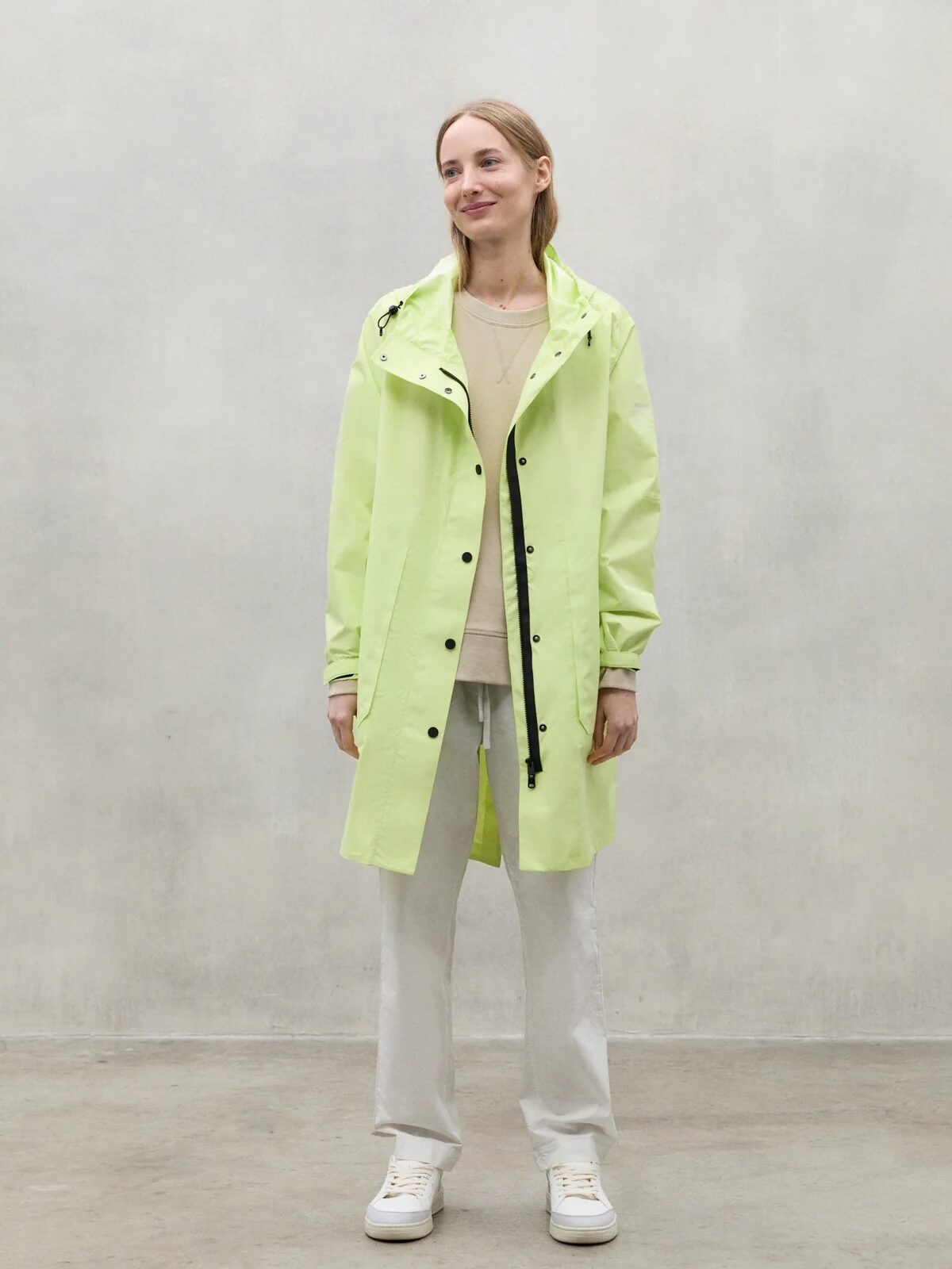A person stands indoors against a plain gray wall, wearing a light yellow waterproof raincoat, white pants, and white sneakers.