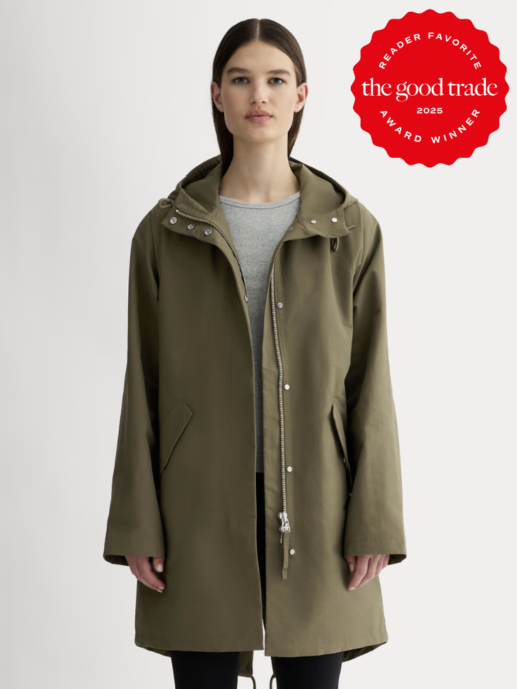 A woman wearing a waterproof olive green hooded raincoat over a gray top stands against a plain background. A red "The Good Trade 2025 Award Winner" badge appears in the top right corner.