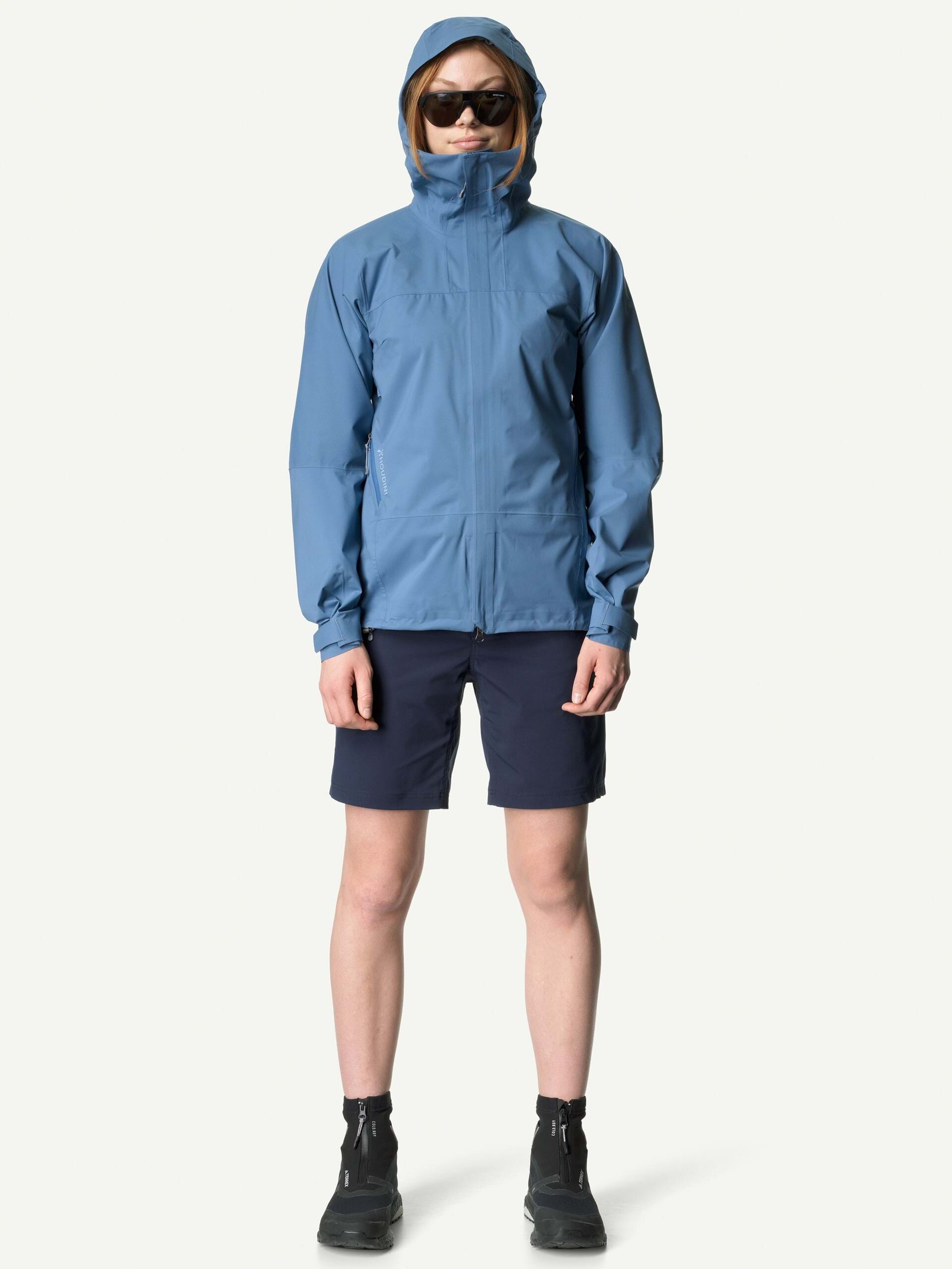 A person wearing a blue waterproof raincoat, navy shorts, black socks, and black shoes stands facing forward against a plain light background.